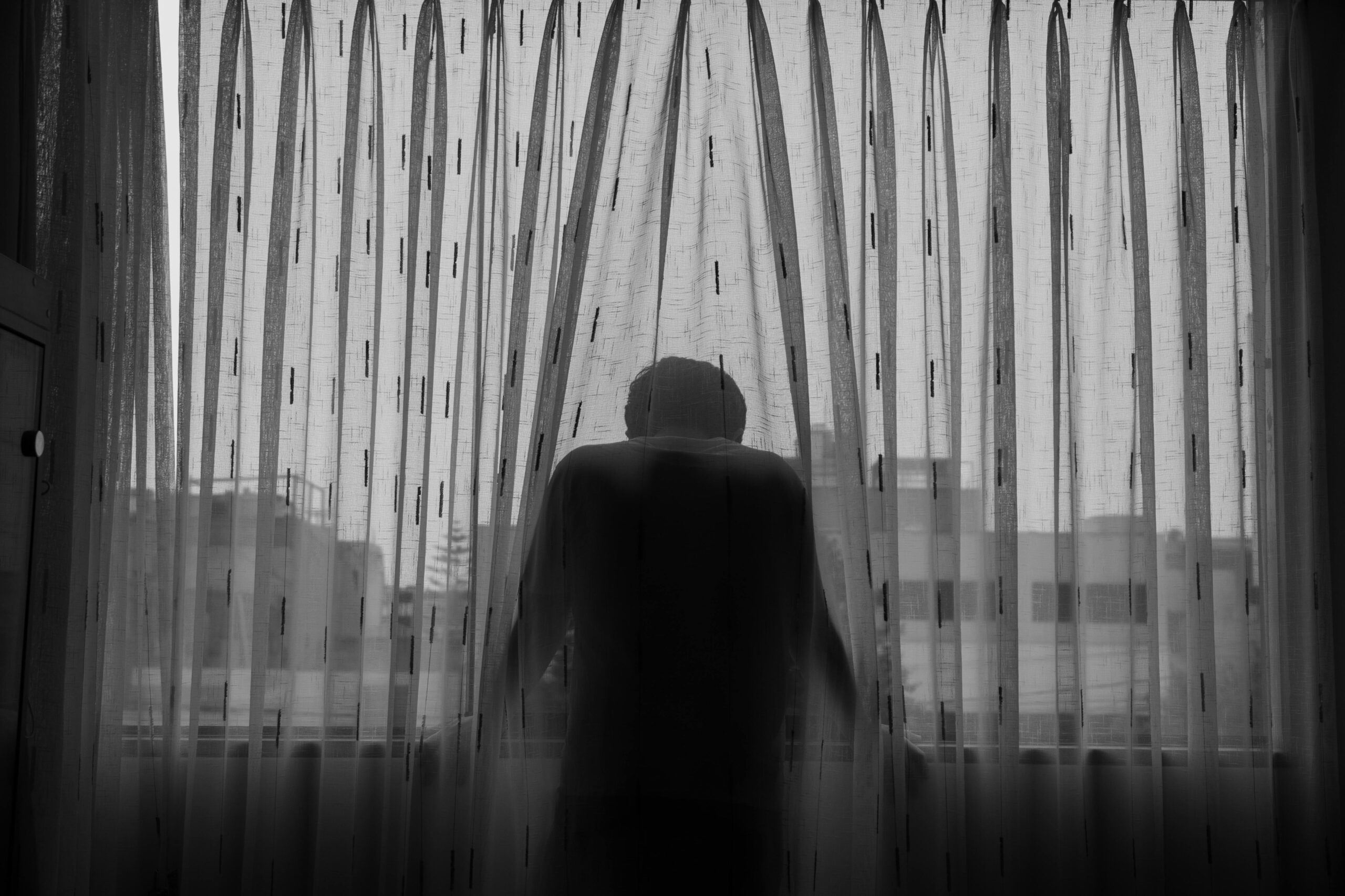 A greyscale photo of a man standing and leaning by the window, covered in sheer curtains.