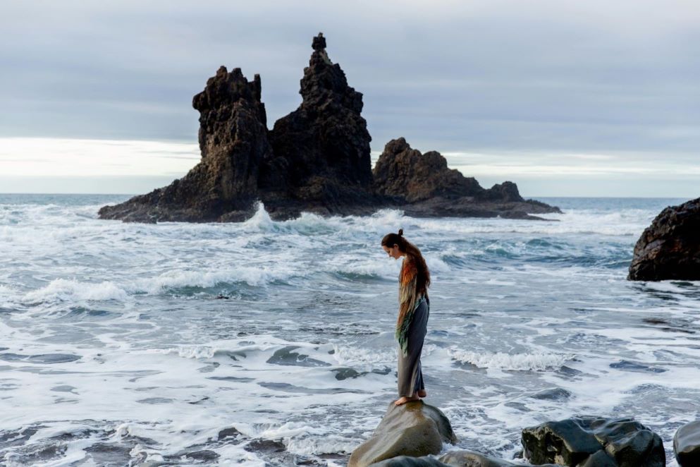A woman standing by the waves on a rock.